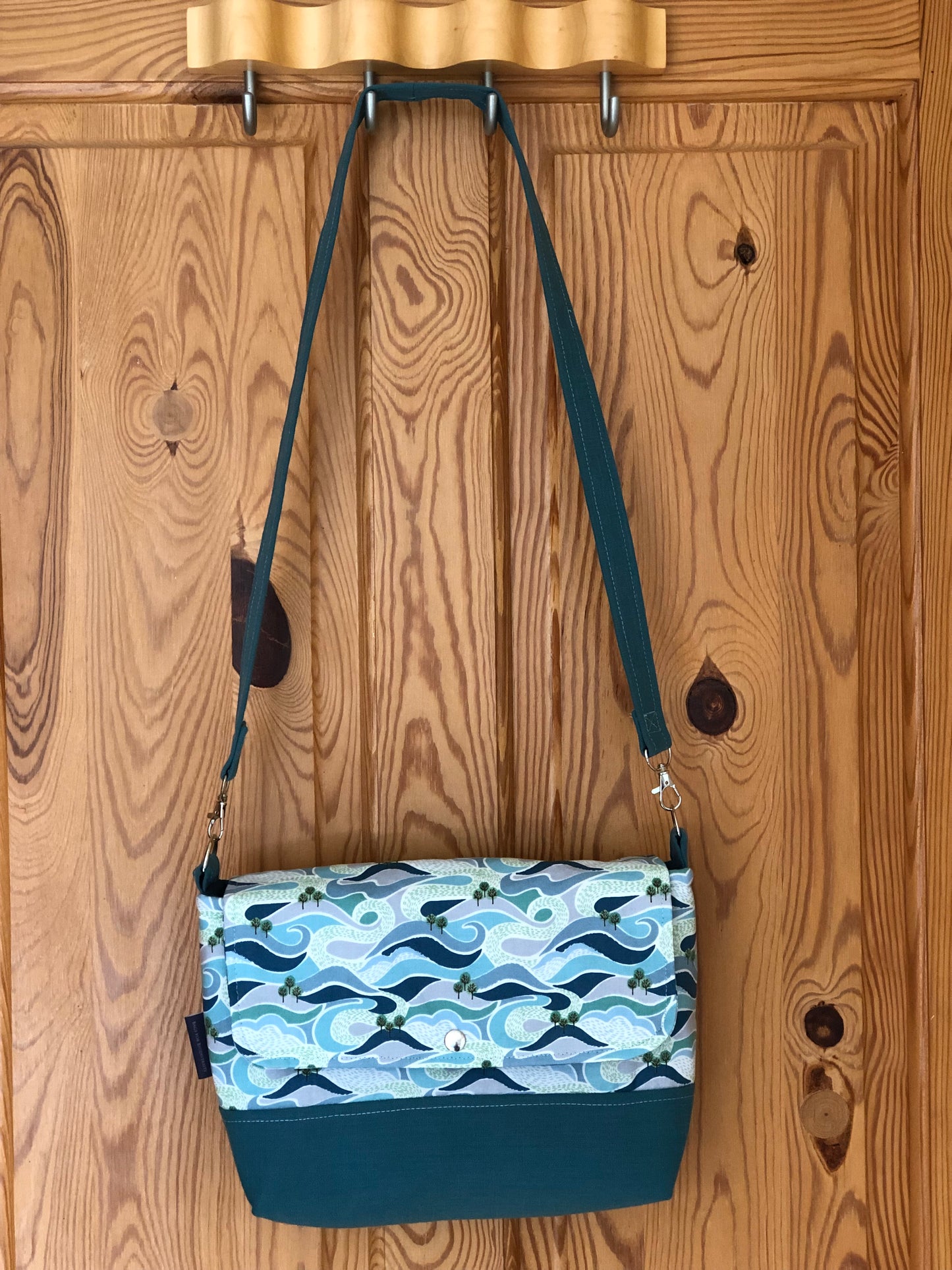 Shoulder bag hanging on back of pine door. The base is turquoise with the body and flap depicting a swirling  green and blue graphic pattern suggesting hills, seas and rivers with groups of trees. There is a stud fastening and detachable handle