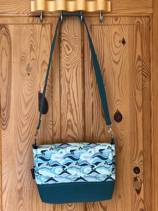 Shoulder bag hanging on back of pine door. The base is turquoise with the body and flap depicting a swirling  green and blue graphic pattern suggesting hills, seas and rivers with groups of trees. There is a stud fastening and detachable handle