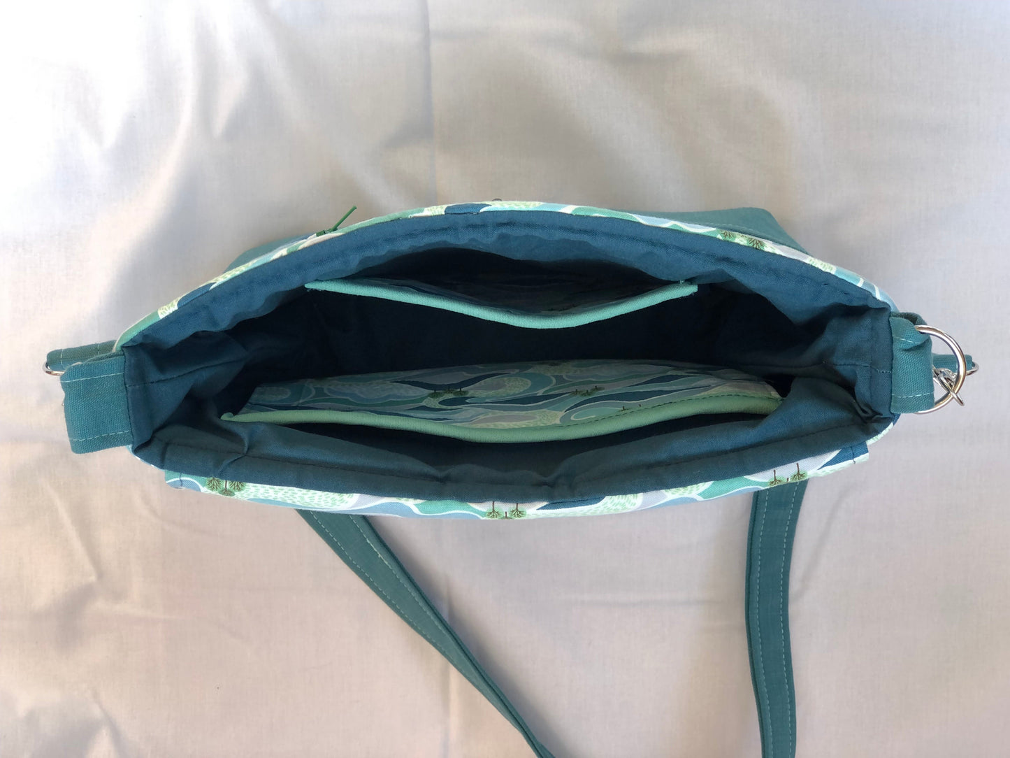 Inside view of shoulder bag, with 2 inside pockets  depicting a swirling pattern of blues and greens with groups of trees. Pockets are bound at the top and the lining is dark turquoise